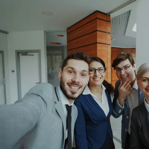 Four diverse business colleagues smiling and taking a selfie in a modern office setting, showing a friendly team spirit.