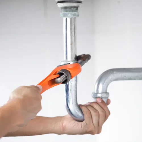 Plumber fixing a metal pipe with an orange wrench under a white tiled sink area.