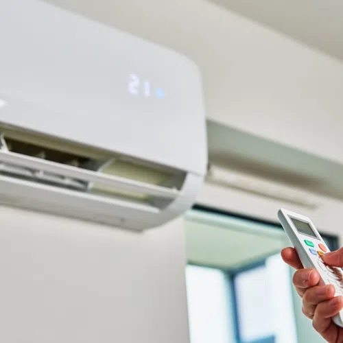 Hand holding remote control pointed at wall-mounted air conditioner in bright room