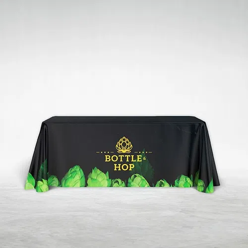 Black tablecloth with green hop illustrations and yellow Bottle & Hop logo on a white floor and wall.