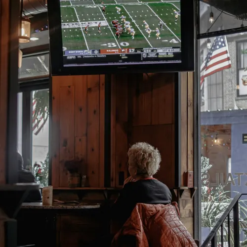 Person watching college football game on TV inside cozy wooden sports bar with American flag outside window
