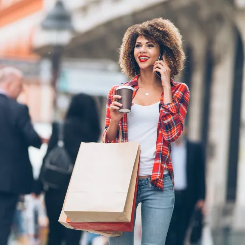 Young woman with curly hair holding shopping bags and coffee cup while talking on phone in city street.