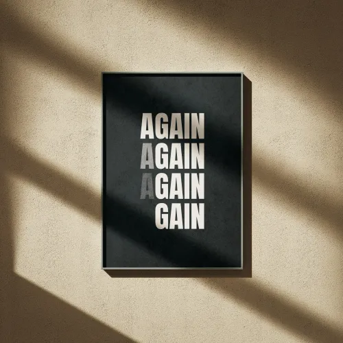 Framed black poster with words AGAIN AGAIN AGAIN GAIN in white and gray letters hanging on textured beige wall.