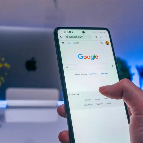 Hand holding smartphone displaying Google search homepage with blurred laptop and plant in background.
