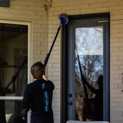 Pest control worker using an extendable brush to clean spider webs off a brick wall near a patio door.