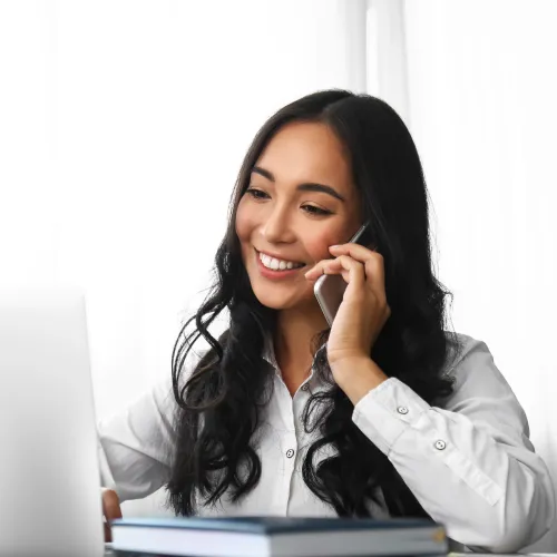 Smiling woman with long black hair talks on phone while working on laptop at bright desk with plant.