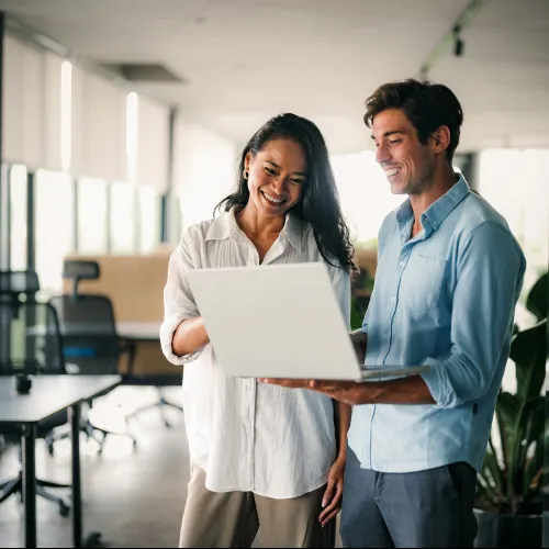 Two colleagues smiling and reviewing content on a laptop in a modern office space with natural light.
