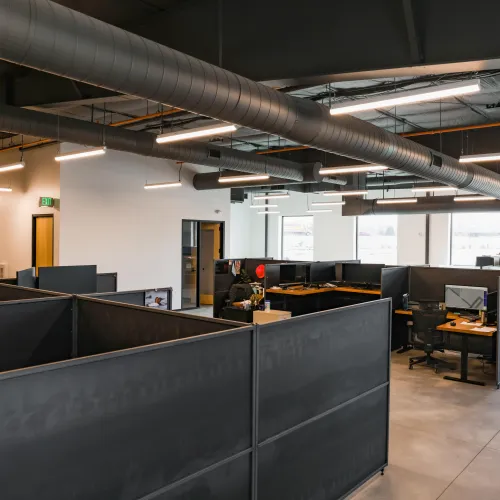 Modern open office space with cubicles, computer workstations, exposed ductwork, and bright overhead lighting.