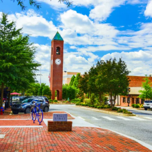 Sunny downtown street with a tall clock tower, brick sidewalks, parked cars, and leafy green trees.
