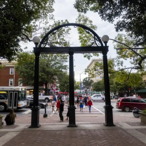 Historic black iron arch at university entrance with students crossing busy city street under trees