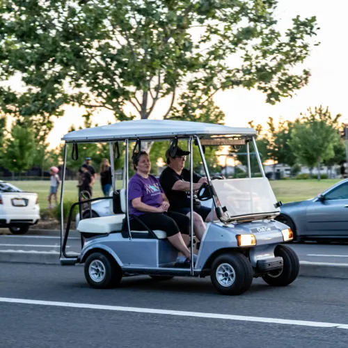 Two people riding in a golf cart on a sunny street with trees in the background.
