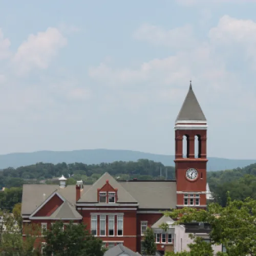 Historic red brick building with clock tower surrounded by trees and distant hills under a partly cloudy sky