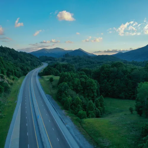 Empty highway winding through lush green hills under a bright blue sky with scattered clouds near mountains.