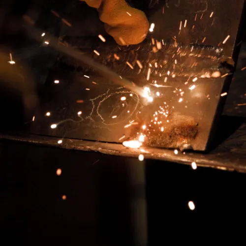 Close-up of welding sparks flying from metalwork with gloved hand holding welding torch in a dark workshop.