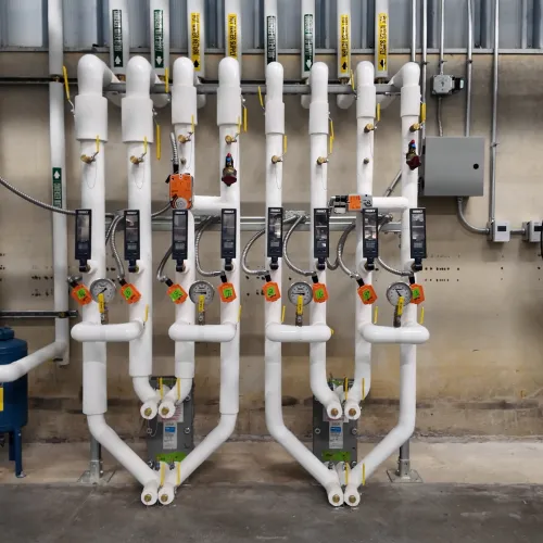 Industrial piping system with multiple white insulated pipes, valves, control units, and large floor fan in a utility room.