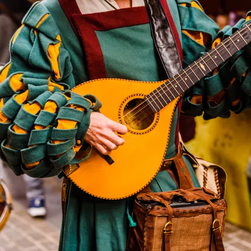 Musician in medieval attire playing a lute with a drummer in the background at an outdoor event