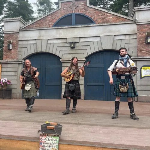 Three musicians in kilts playing string instruments on a wooden stage in front of a brick and stone building.