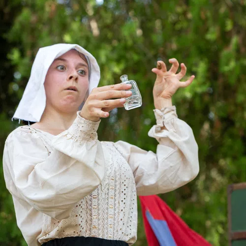 Woman in historical costume holding a small glass bottle with a surprised expression, outdoors with greenery behind.