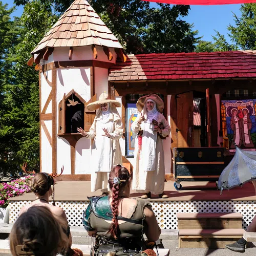 Performers in white robes acting on stage in front of a medieval-themed set with audience watching outdoors.