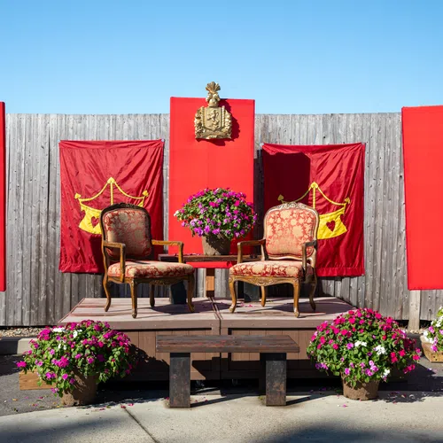 Outdoor wooden stage with vintage chairs, red banners, potted flowers, and a crowned emblem under clear blue sky.