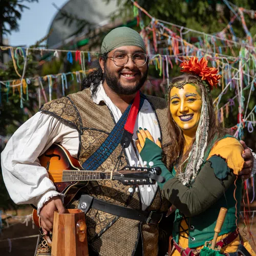 Smiling man in medieval garb holding a mandolin and woman in colorful costume with yellow face paint outdoors