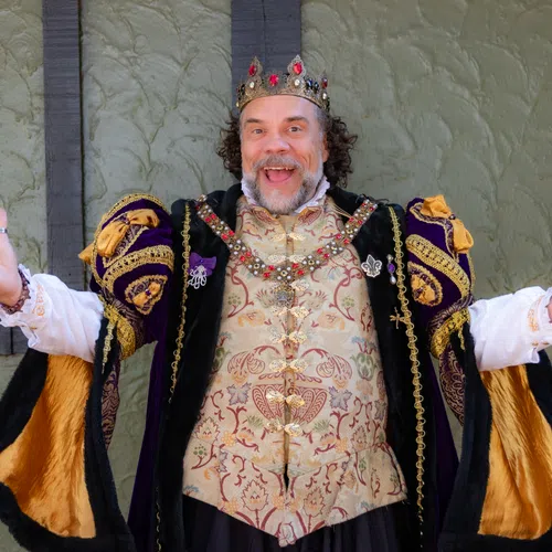 Man dressed as a king in ornate medieval robe and crown, smiling with arms open wide indoors