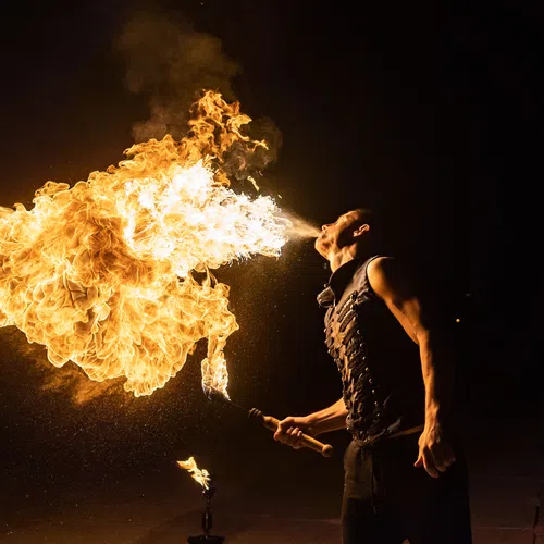 Fire performer blows a large flame into the night holding a torch with muscular arms visible.