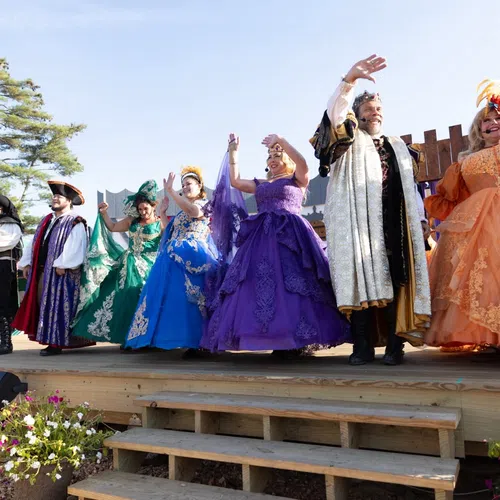 Performers in colorful Renaissance costumes wave from a wooden stage outdoors with trees and blue sky behind them.