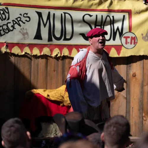 Performer in red cap and tattered clothing acts at Sturdy Beggar's Mud Show in front of wooden backdrop and crowd