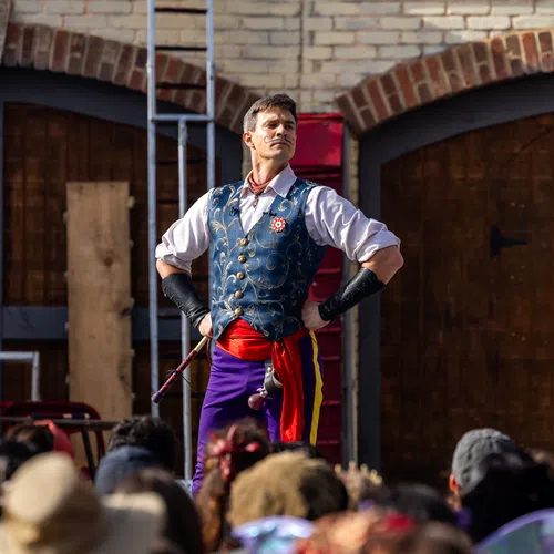 Performer in colorful costume with hands on hips stands confidently before a crowd in front of a brick wall.