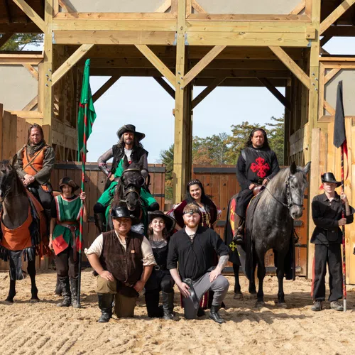 Group of medieval reenactors in authentic costumes with horses standing in front of a wooden fortress.