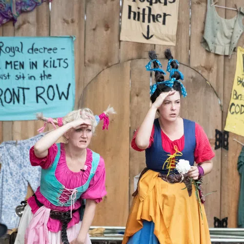 Two women in colorful Renaissance costumes with unique hairstyles performing on a wooden stage with signs behind them