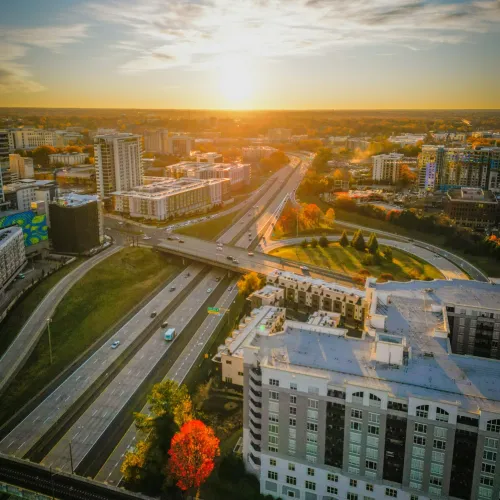 Aerial view of city highways, buildings, and autumn trees at sunset with golden sky and urban landscape.