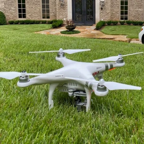 White quadcopter drone with camera landing on green grass in front of a brick house with black door.
