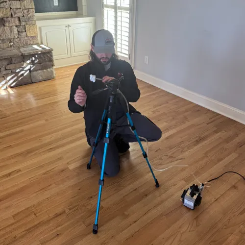 Photographer kneeling on hardwood floor setting up camera on tripod inside bright living room with stone fireplace.