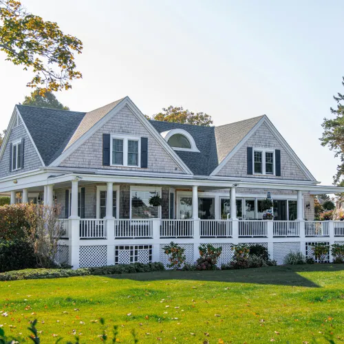 Large white cottage with wraparound porch surrounded by green lawn and trees on a clear day