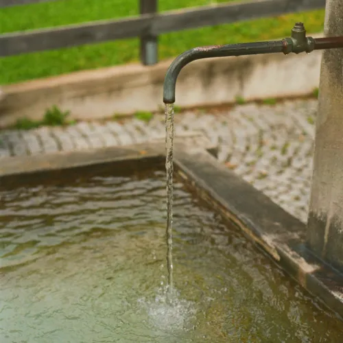 Outdoor metal tap pouring water into a stone basin with a green grass background and wooden fence.