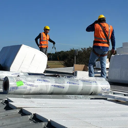 Two construction workers in safety gear installing insulation panels on a commercial metal roof under clear sky.