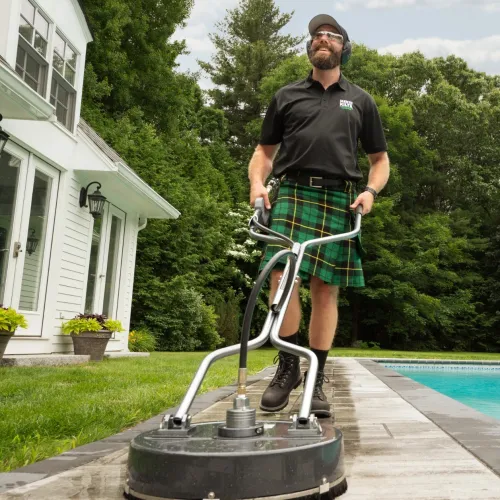 Man in a green kilt power washing a stone patio near a pool beside a white house on a sunny day.