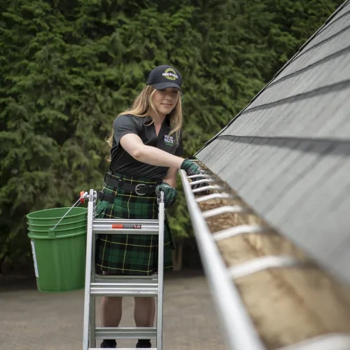 Young woman wearing gloves and cap cleans leaves from a home's gutter while standing on a ladder outdoors.