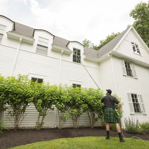 Man in kilt cleaning second-story windows of white house with long pole washer on sunny day