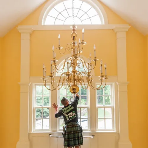 Man in a kilt stands on a ladder cleaning a large gold chandelier in a room with yellow walls and arched windows