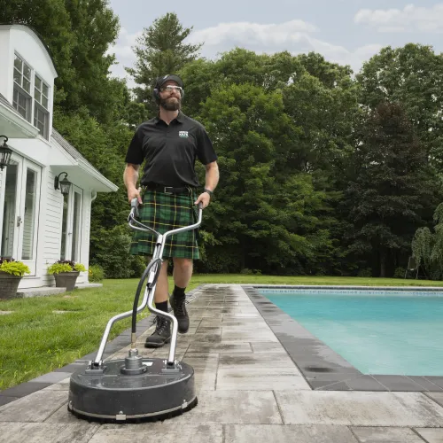 Man in kilt cleaning stone patio near swimming pool with surface cleaner machine on a sunny day.