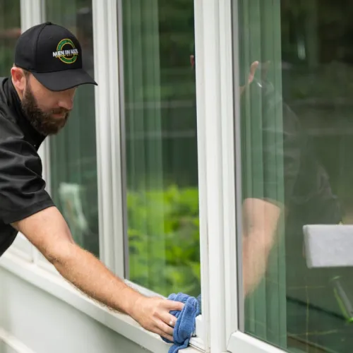 Man in black shirt and cap cleaning white-framed windows with blue cloth outdoors during daytime.