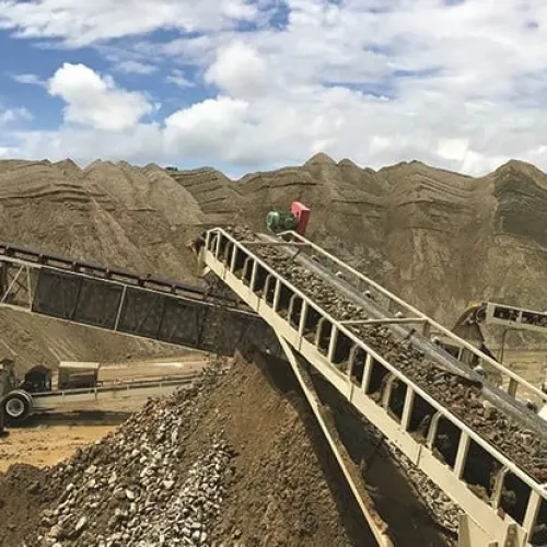 Conveyor belts transporting crushed stones and gravel at an open mining site with large earth piles and blue sky