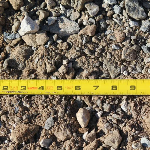Yellow measuring tape stretched over gravel and dirt showing measurements up to 11 inches.