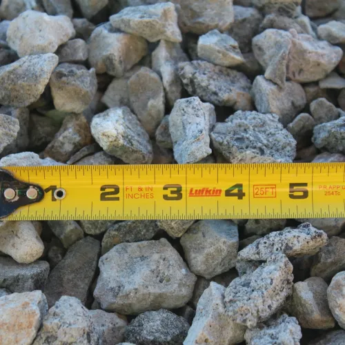Yellow tape measure laid over a pile of gray gravel rocks measuring size in inches.