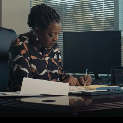 Woman writing notes at desk with computer and documents in modern office with window blinds.