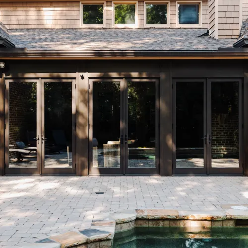 Modern home exterior with large glass doors opening to a stone patio and outdoor pool area.
