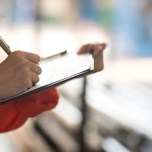 Person in orange sleeve writing on clipboard with blurred background of benches and sunlight.